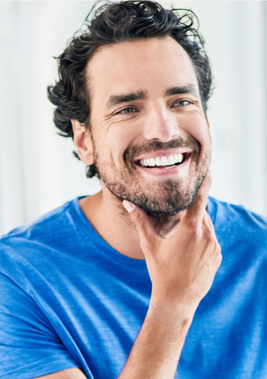 Smiling man touching his beard in blue shirt.